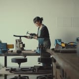 파란색 바이스들이 놓인 작업대가 있는 작업실. 가운데에 한 사람이 서서 장갑 낀 손으로 망치를 들고, 바이스에 고정된 막대 위에 판을 대고 있는 모습.