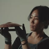 밝은 회색 벽 앞. 왼쪽에서 장갑 낀 두 손이 구릿빛 금속 조각을 받쳐 들고, 오른쪽의 인물이 그 조각을 바라보는 모습.