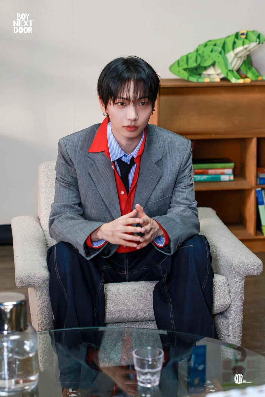 A BOYNEXTDOOR member in a grey blazer and red shirt, seated in a cream armchair with his hands steepled