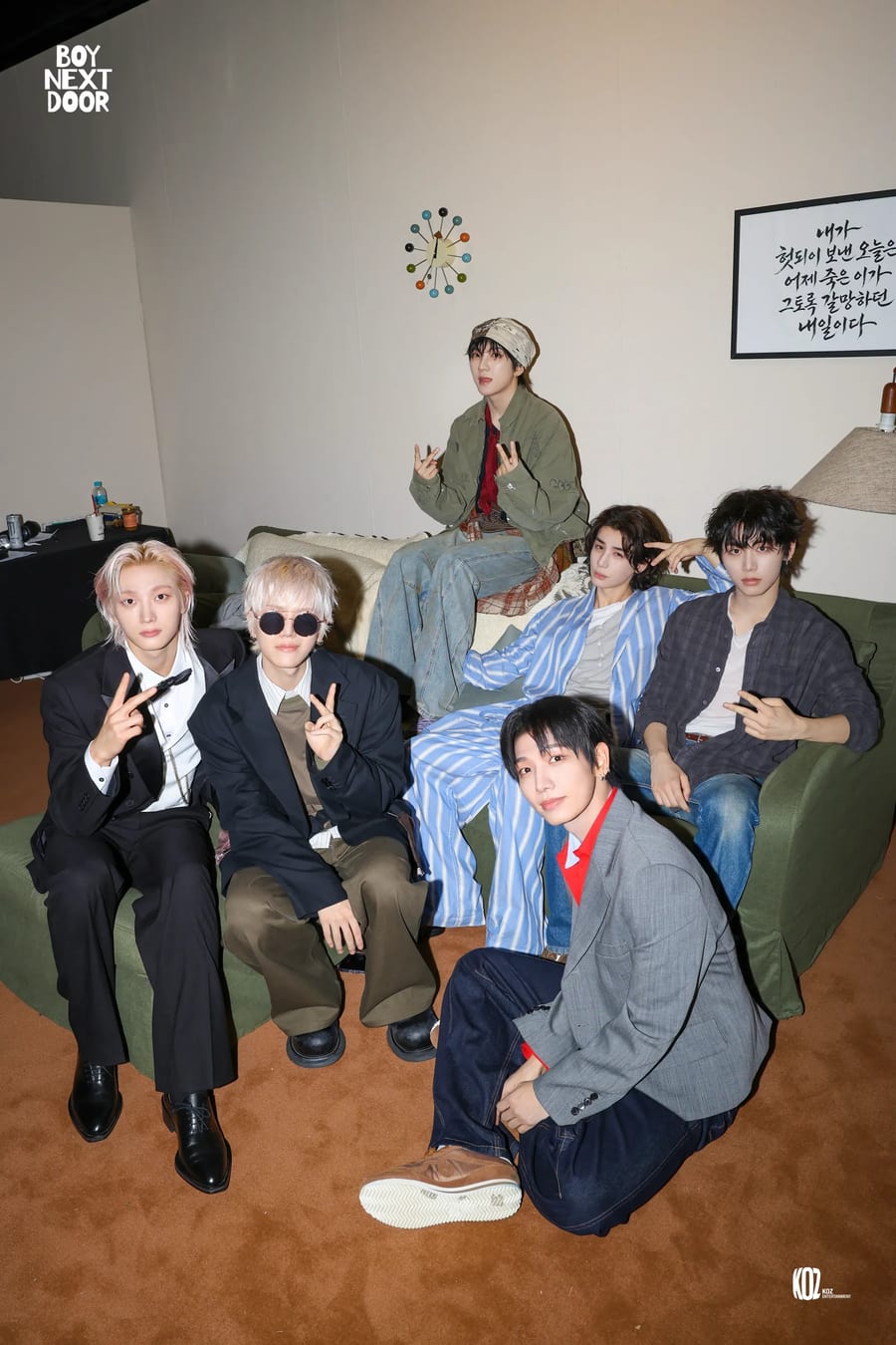 BOYNEXTDOOR on and around a green sofa in a set living room, under a wall clock and a framed Korean calligraphy piece