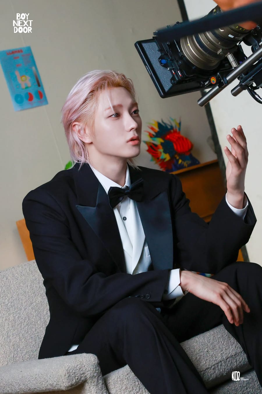 A BOYNEXTDOOR member with pink hair in a black tuxedo and bow tie, seated beside a cinema camera rig